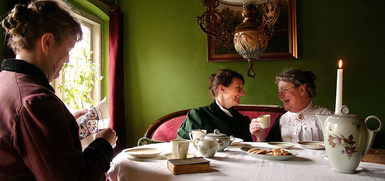 1910 : Sønderjysk kaffebord i Als hvor Lærerinde Frøken Ludvigsen (tv) er hjemvendt fra Aabenraa og beretter nyt til Fyrbøderfrue Møller og Boghandlerfrue Johansen om kvindernes emancipering (frigørelse og ligestilling). På grund af tyskernes belejring må der kun undervises i tysk i skolerne i det sønderjyske, ligesom danske sange ikke måtte kunne høres udenfor. Ved kaffebordet nyder man derfor jødekager der er bagt i det nymodens støbejernskomfur og slutter hyggen med en dansk sang fra højskolesangbogen. Lærerinde Frøken Ludvigsen underviser i dansk historie og dansk sprog privat. Fyrbøderfrue Møller underviser i klaver i hjemmet imens Boghandlerfruen står for husførelsen hjemme.